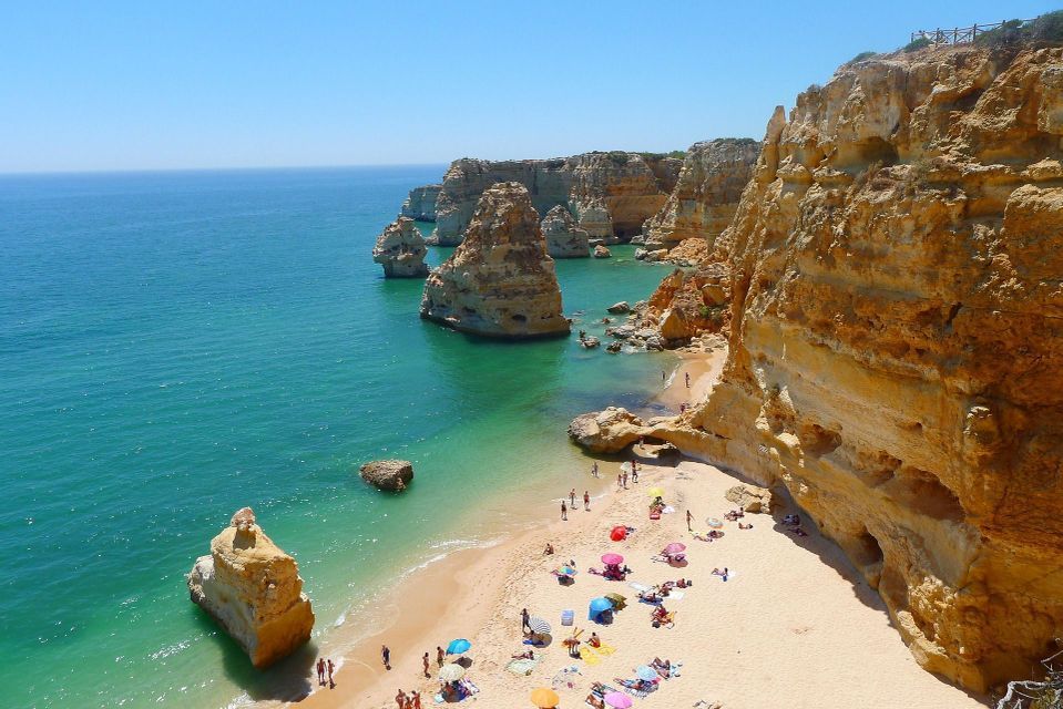 Vista dall'alto di una cala sabbiosa affollata di persone, circondata da alte scogliere dorate e mare turchese.