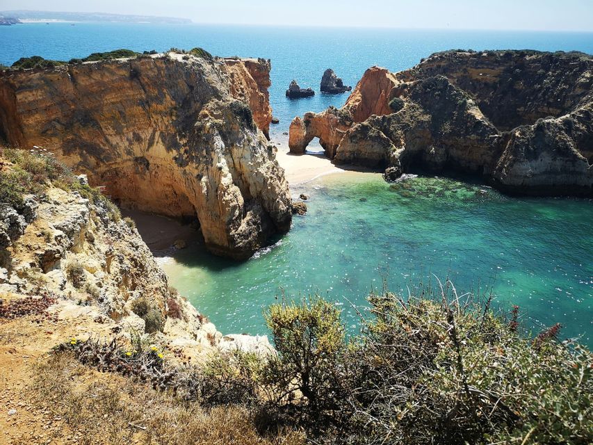 Vista soleggiata di una cala costiera con grandi formazioni rocciose, un arco naturale sulla spiaggia e acqua turchese.