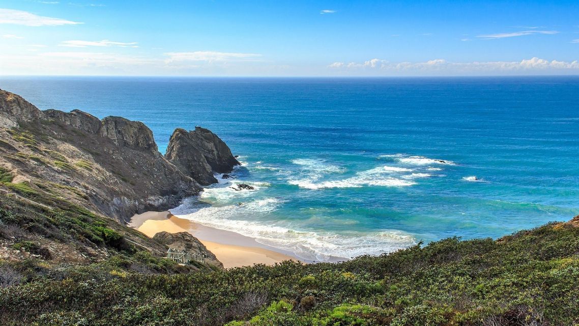 Panorama mozzafiato da una verde scogliera che domina una spiaggia sabbiosa isolata, formazioni rocciose e un vasto oceano blu sotto un cielo limpido.