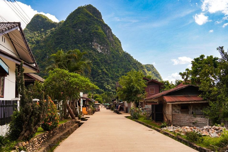 Une rue paisible traverse un village champêtre, bordée de maisons et d'arbres, menant vers une grande montagne boisée sous un ciel bleu.