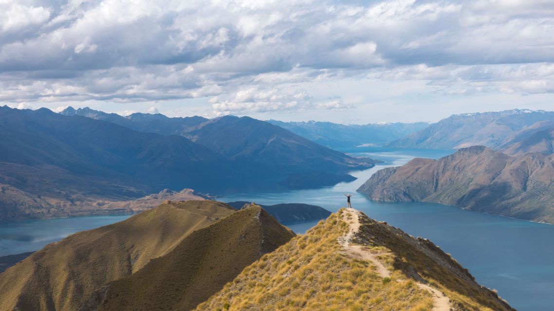 Eine Person steht mit erhobenen Armen auf einem Berggipfel und blickt auf eine weite Landschaft aus Bergen und einen sich schlängelnden See.