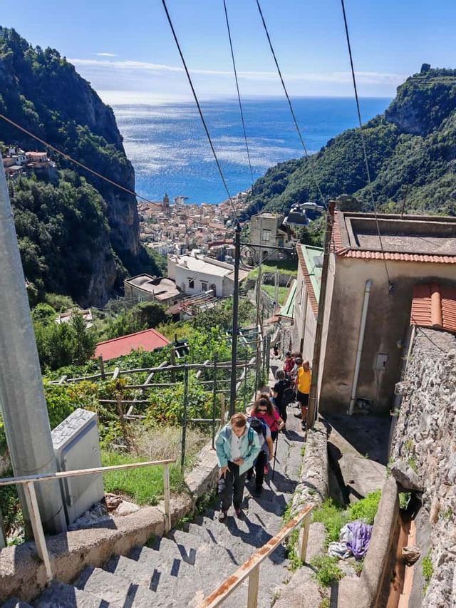 Un viaje en grupo de WeRoad sube una larga escalera de piedra con vistas a un pueblo costero enclavado entre colinas verdes y el mar.