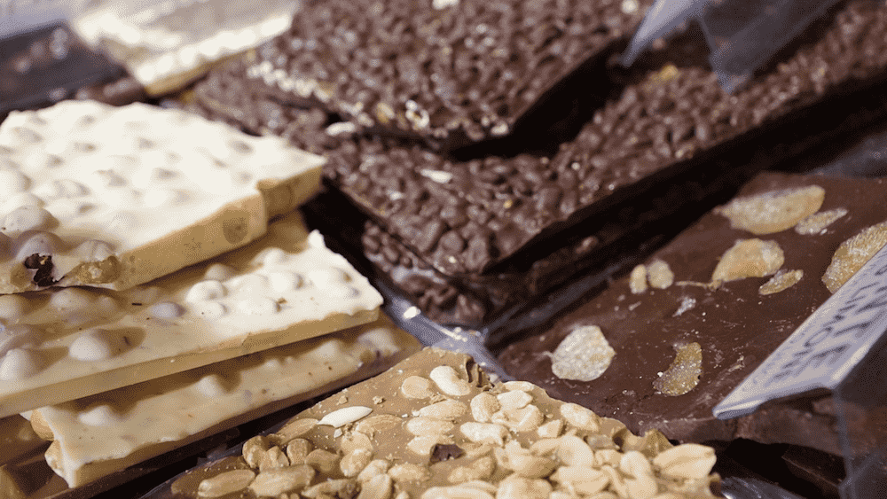 Un assortimento in primo piano di scaglie di cioccolato artigianali, tra cui fondente, bianco e al latte, con varie guarnizioni di frutta secca e frutta.