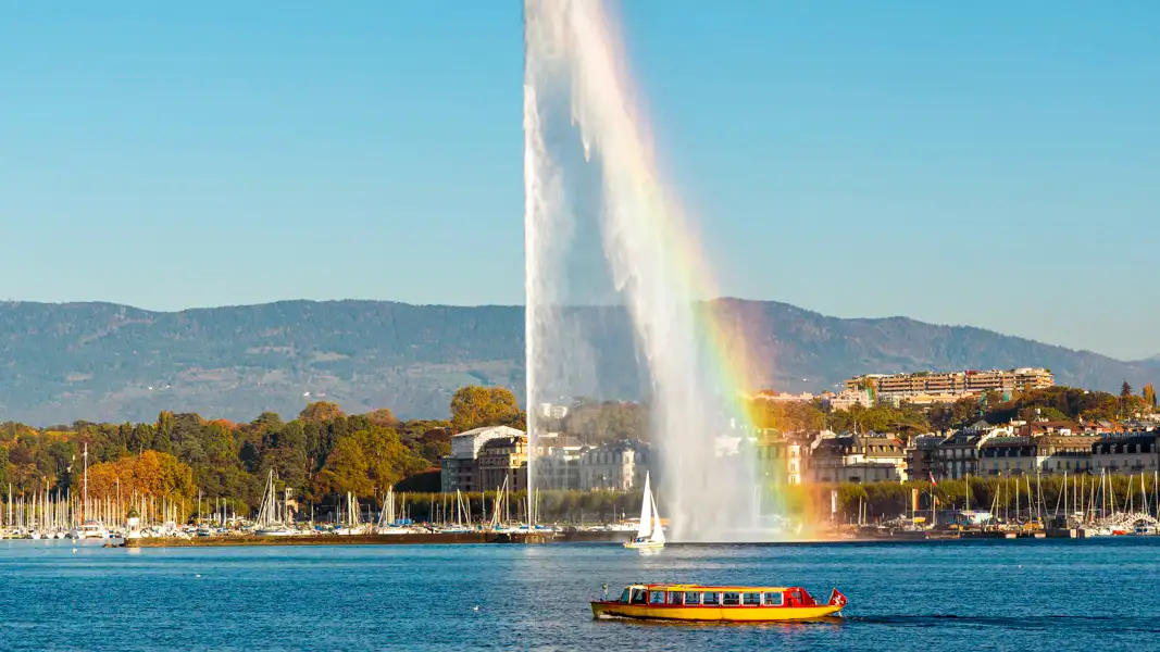 A large fountain on a lake creates a rainbow in its spray as a yellow boat passes, with a city and mountains behind.