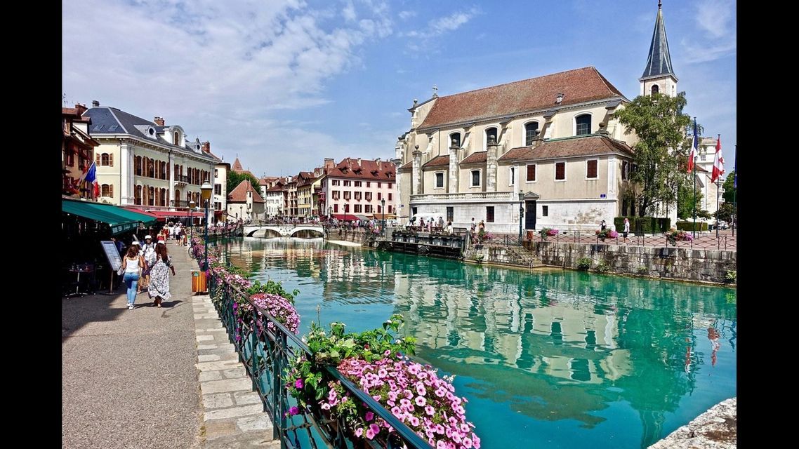 Un canale turchese scorre attraverso una città storica con persone che passeggiano lungo il lungomare, oltre vecchi edifici e una chiesa.