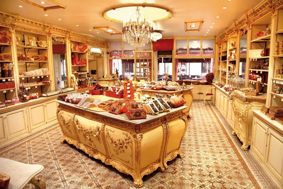 L'interno sfarzoso di una pasticceria, con un lampadario di cristallo e ampi banconi che espongono cioccolatini e dolci assortiti.