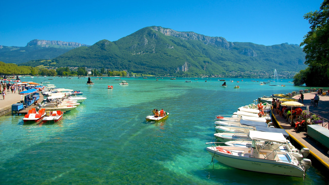 Numerous boats are docked and floating on a clear turquoise lake, with lush green mountains rising in the background under a blue sky.