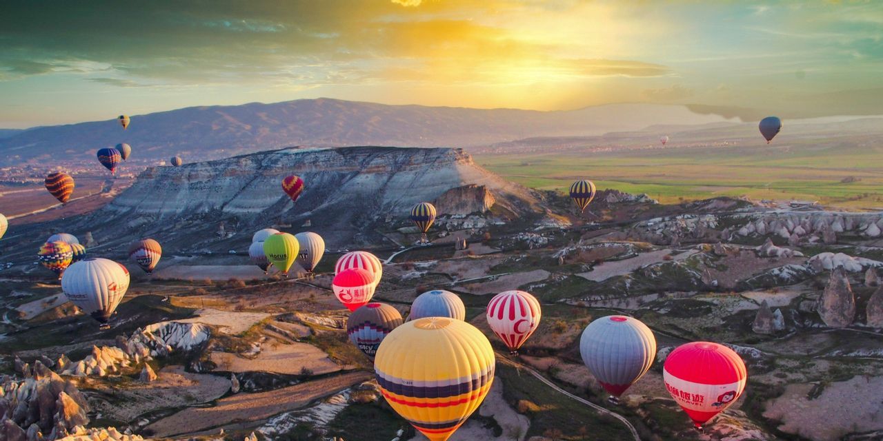 Dozens of colorful hot air balloons float over a vast, rocky landscape with distant mountains during sunrise.