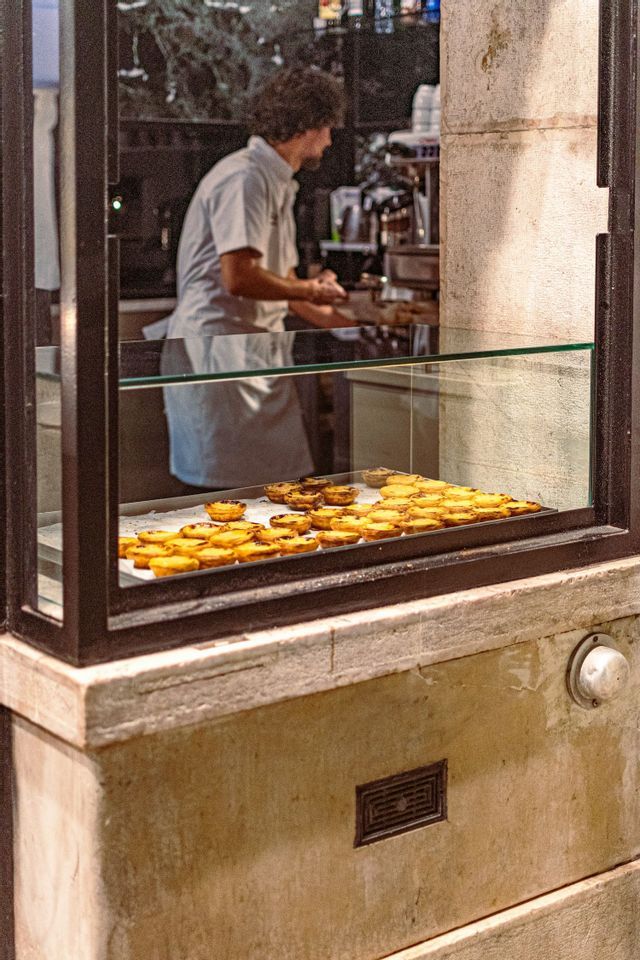 Una vetrina di una pasticceria piena di pastéis de nata, con un fornaio al lavoro sullo sfondo.