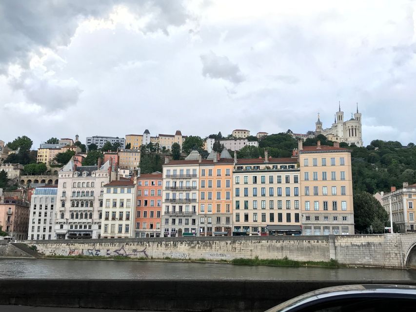 Des bâtiments colorés bordent une rive devant une colline verte coiffée d'une grande cathédrale sous un ciel nuageux.