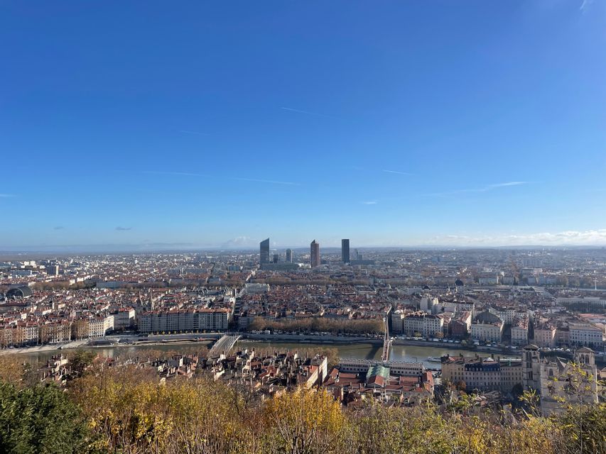 Une vue panoramique d'une vaste ville traversée par une rivière et des ponts, prise depuis une colline parée de feuillage d'automne sous un ciel bleu dégagé.