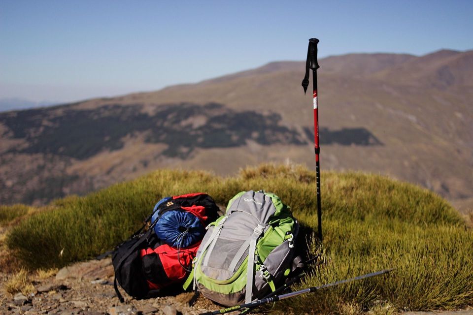 Dos mochilas de senderismo y bastones de trekking descansan en una ladera montañosa cubierta de hierba en un día soleado.