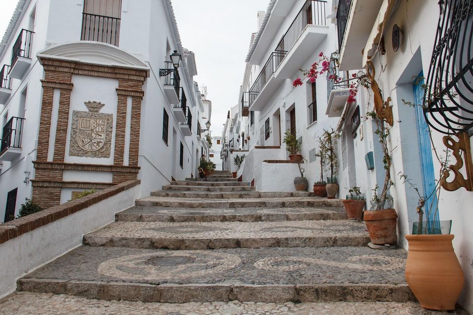 Una escalera de piedra sube por una calle estrecha entre edificios encalados con balcones y un escudo de piedra.