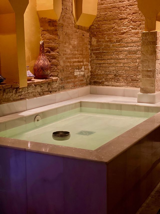 Una piscina cubierta revestida de mármol en una sala con paredes de ladrillo visto y arcos arquitectónicos amarillos.