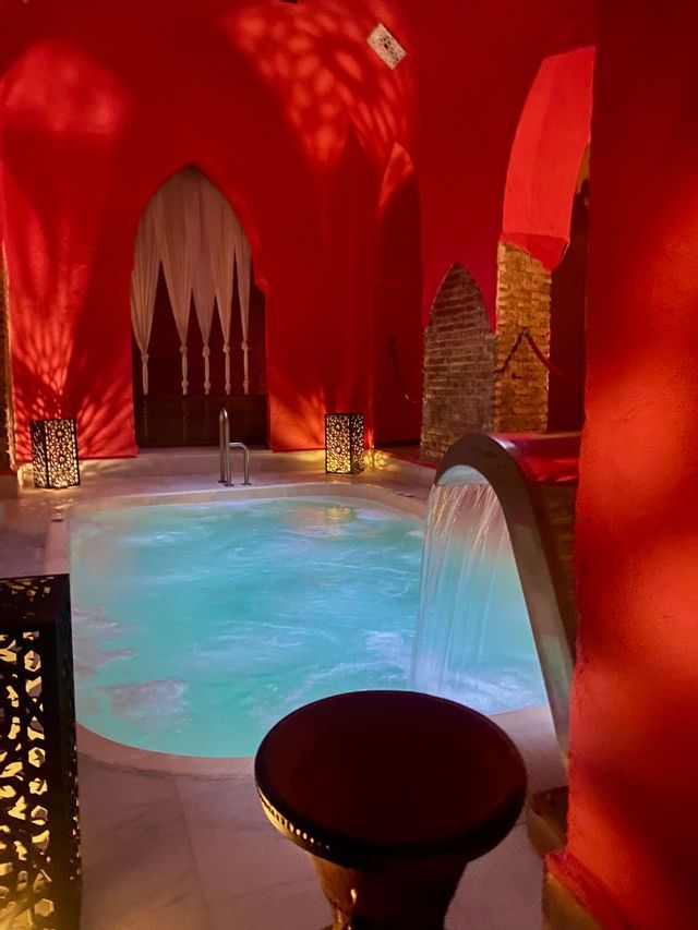 Una piscina interior turquesa con cascada dentro de un spa con paredes rojas, puertas arqueadas y faroles decorativos.