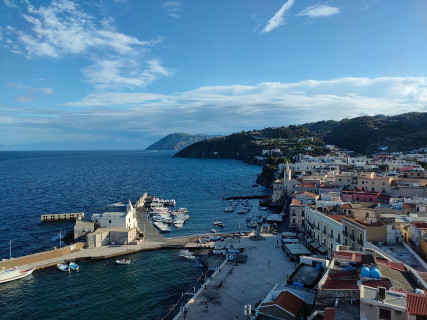 Una vista dall'alto di una città costiera con un porto pieno di barche, un lungo molo e una costa collinare sotto un cielo parzialmente nuvoloso.