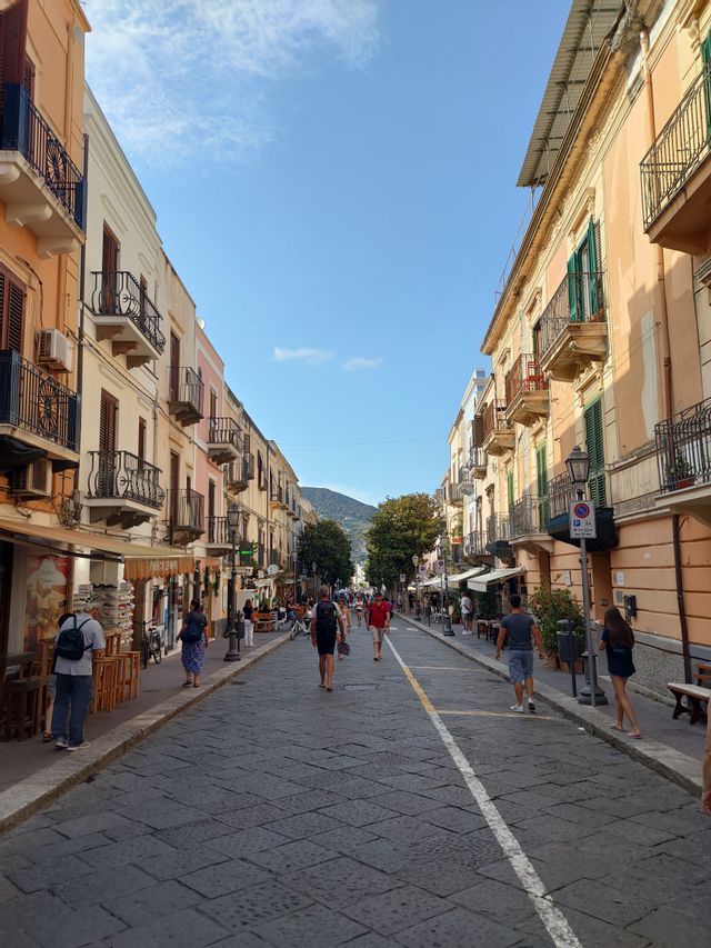 Gente che passeggia lungo una stretta via acciottolata, fiancheggiata da edifici colorati con balconi, sotto un limpido cielo azzurro.