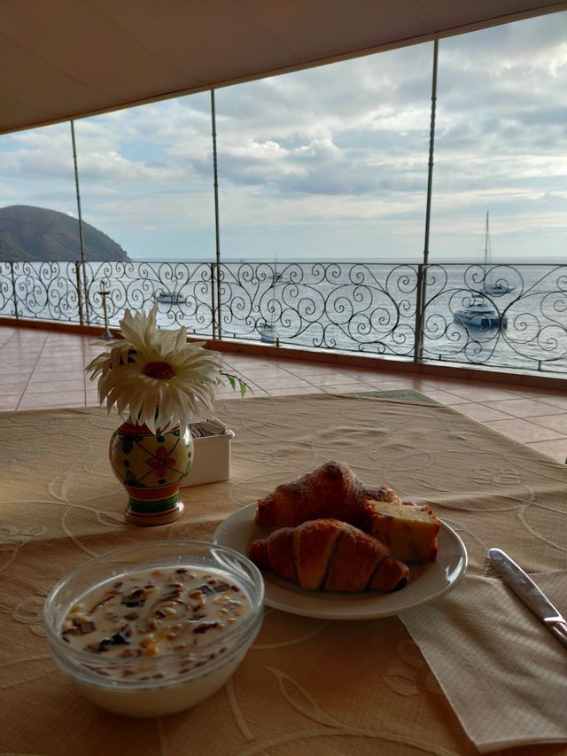 Una colazione di cornetti e cereali su un tavolo con vista su un mare calmo con diverse barche, da una terrazza.