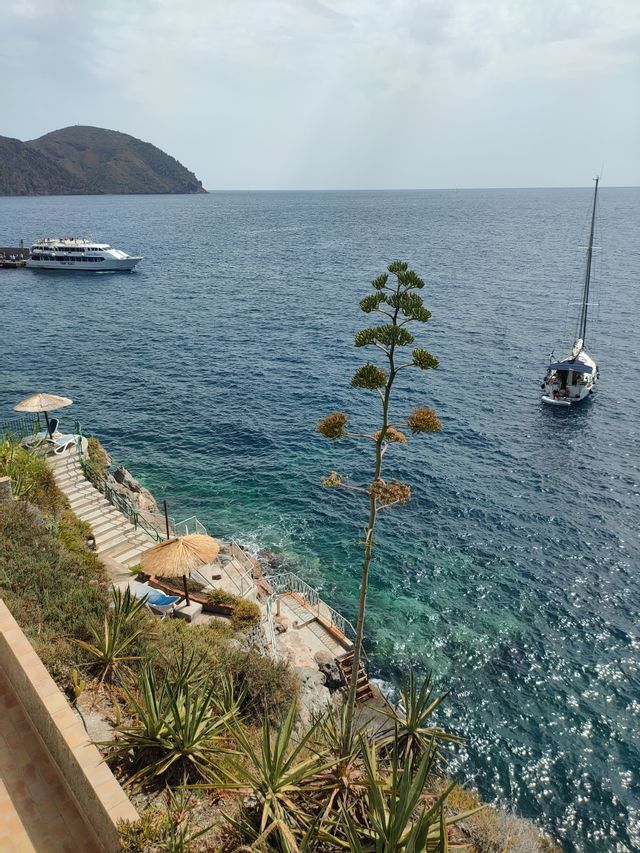 Vista dall'alto di una costa rocciosa con scalinata che scende al mare blu cristallino, dove sono ormeggiati una barca a vela e un traghetto.