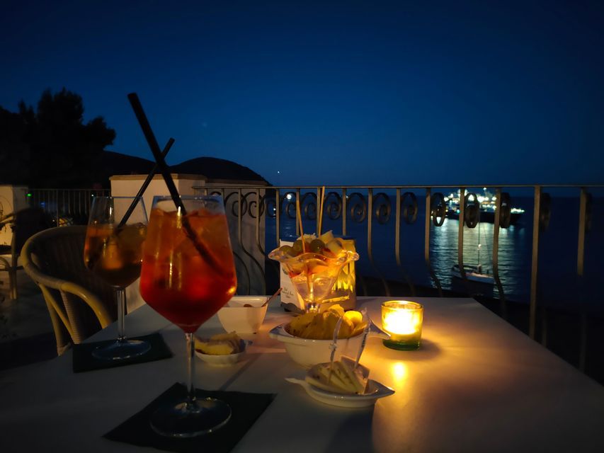 Due cocktail all'arancia e stuzzichini su un tavolo illuminato da una candela su una terrazza con vista mare di notte.