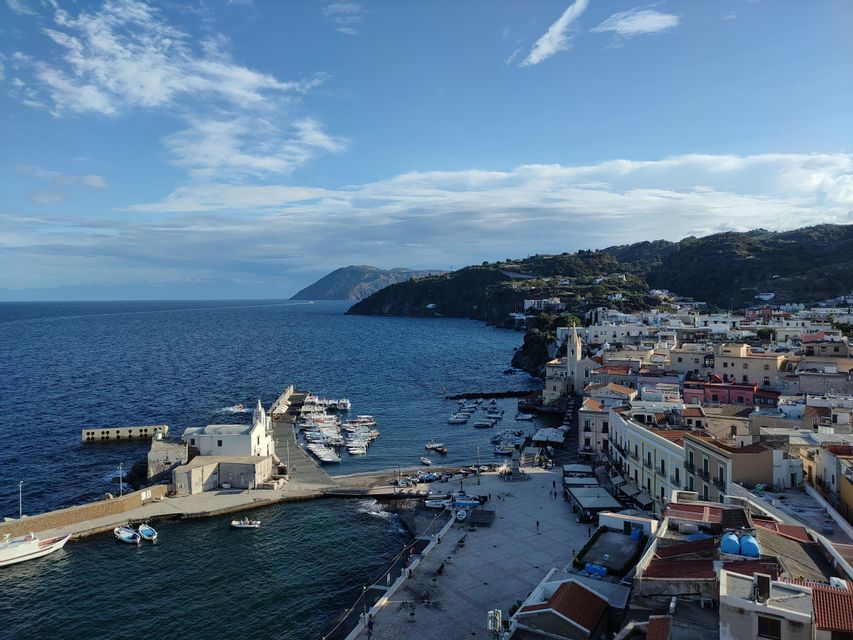 Vista aerea di una colorata cittadina costiera e del suo porto pieno di barche, adagiata su un litorale collinare sotto un cielo azzurro.