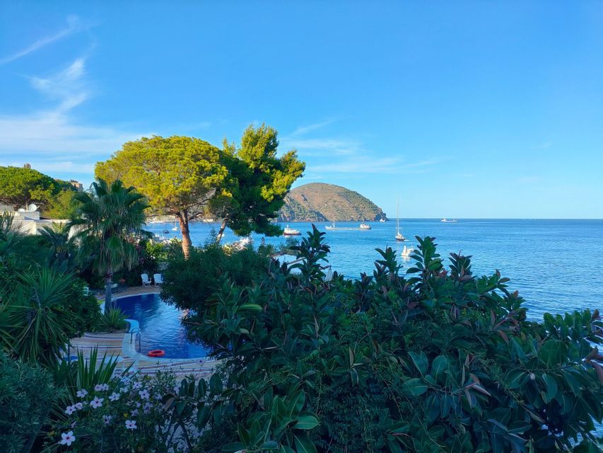 Una piscina circondata da alberi rigogliosi si affaccia su un mare tranquillo costellato di barche e un'isola collinare in lontananza.