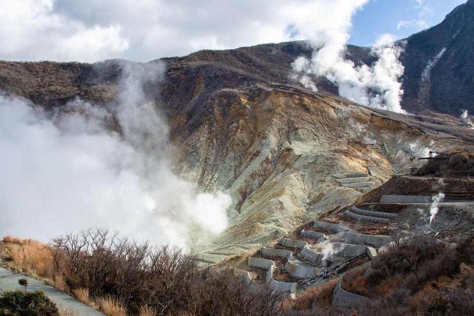 Vapore e fumo si innalzano da sfiatatoi geotermici su un versante montuoso roccioso e terrazzato sotto un cielo nuvoloso.