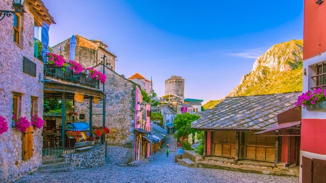 Eine verwinkelte Kopfsteinpflastergasse, gesäumt von farbenfrohen Steingebäuden und Balkonen mit rosa Blumen in einer historischen Stadt.