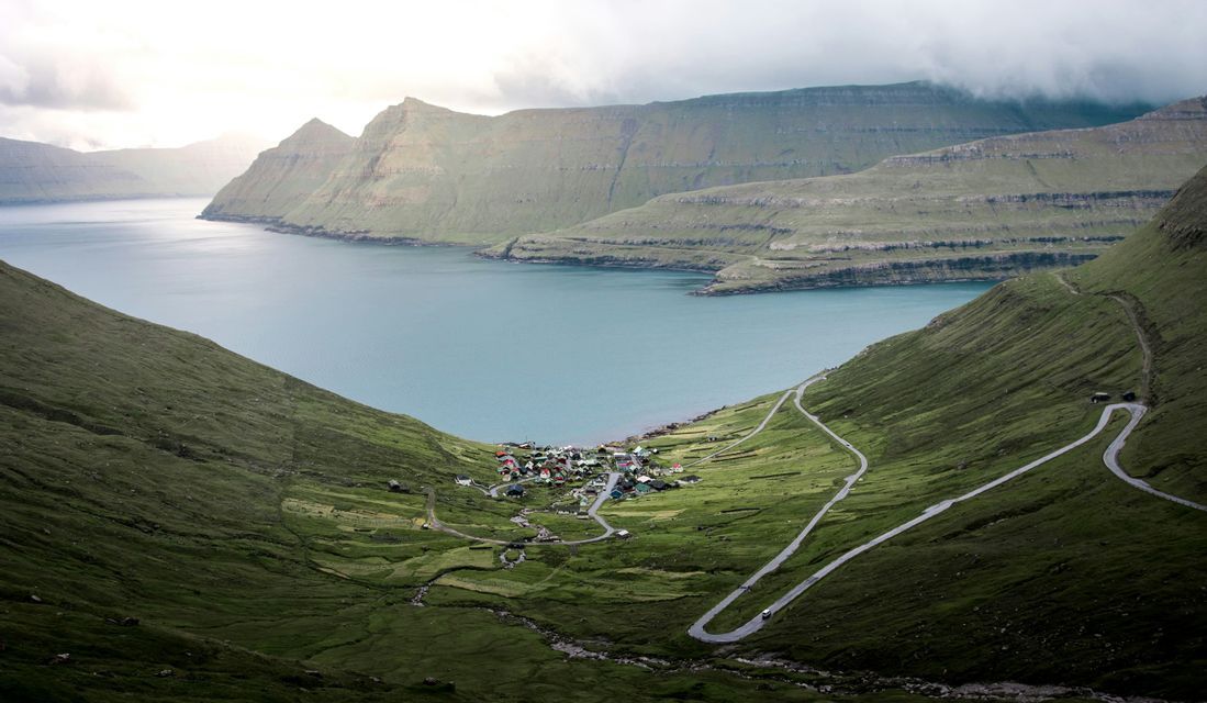 Un petit village aux toits colorés est situé sur la rive d'un fjord, entouré de montagnes vertes escarpées et d'une route sinueuse.