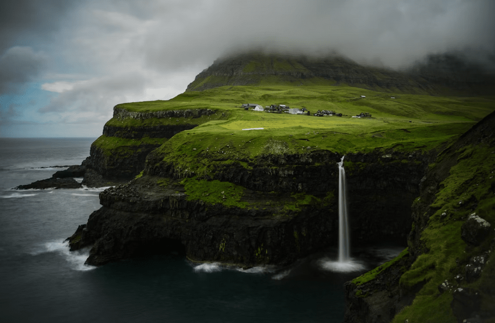 Une cascade dévale d'une falaise verdoyante directement dans l'océan, avec un petit village et une montagne brumeuse en arrière-plan.