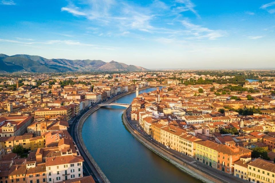 Luftaufnahme einer Stadt mit terrakottafarbenen Dächern, die sich entlang eines breiten, gewundenen Flusses erstreckt, im Hintergrund ferne Berge unter blauem Himmel.