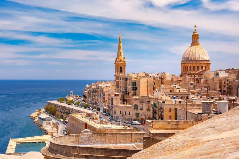 A coastal city with a church dome and spire overlooks the blue sea under a partly cloudy sky.