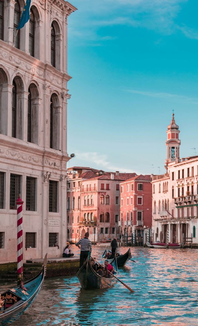 Góndolas con gente flotando por un canal junto a edificios históricos bajo un cielo azul claro.