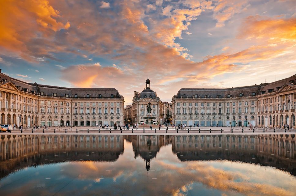 Un grand bâtiment historique se reflète parfaitement dans un miroir d'eau sur une place de ville sous un ciel de coucher de soleil coloré.