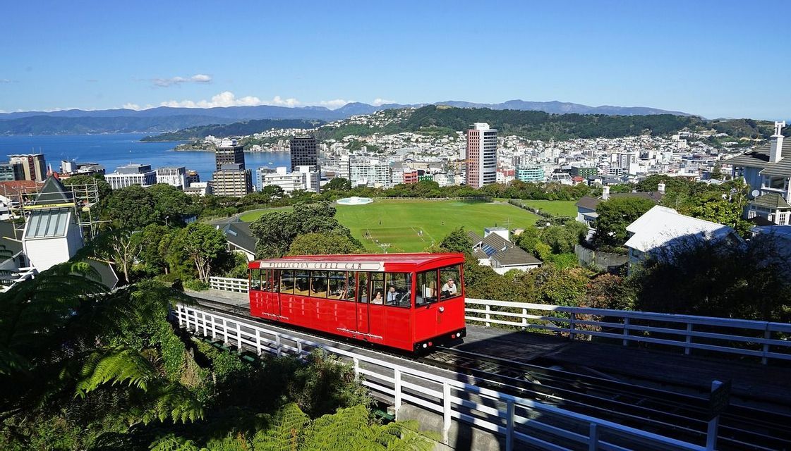 Eine leuchtend rote Seilbahn mit Fahrgästen fährt auf einer Strecke mit Blick auf eine Küstenstadt mit einem großen grünen Park und fernen Bergen.