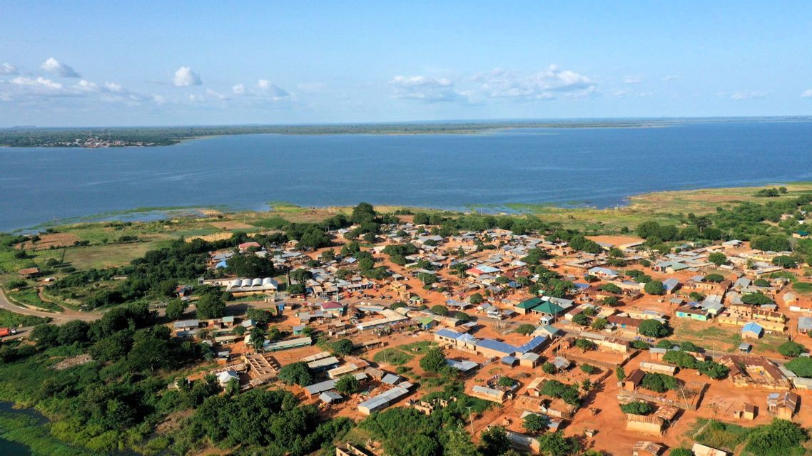 Luftaufnahme eines kleinen Dorfes mit unbefestigten Wegen und verstreuten Bäumen, gelegen am Ufer eines großen Gewässers unter blauem Himmel.