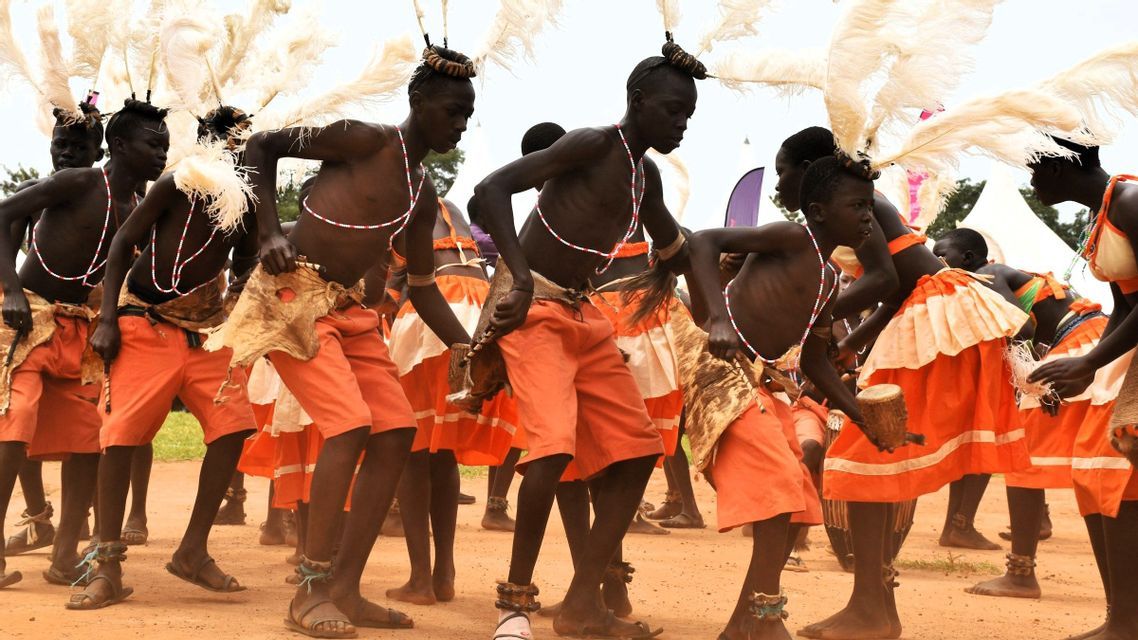 Eine Gruppe von Menschen in orangefarbenen Trachten und weißen Federkopfschmuck führt einen kulturellen Tanz im Freien auf unbefestigtem Boden auf.