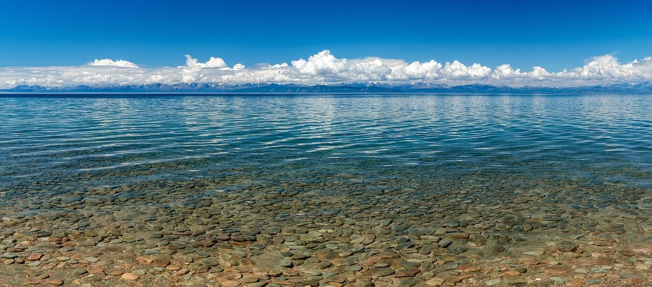Una vista panoramica di un lago limpido con fondale roccioso, montagne innevate in lontananza e un cielo azzurro brillante con nuvole.