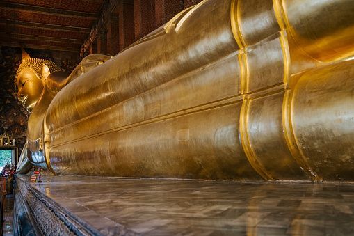 A low-angle view of a large, golden reclining Buddha statue resting on a marble platform inside an ornate temple.