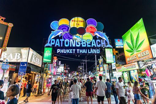 A crowd of people walking down a busy street at night, illuminated by a large colorful neon archway and various shop signs.