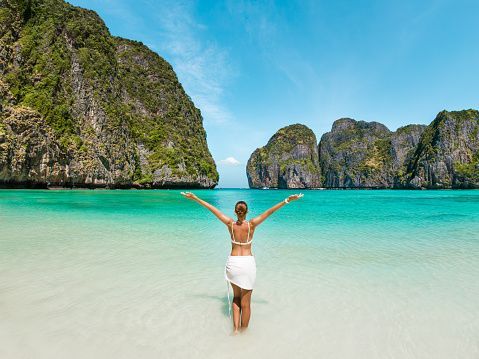 A woman seen from the back, wearing a white bikini and sarong, stands with arms outstretched in shallow turquoise water, facing large green cliffs across the bay.