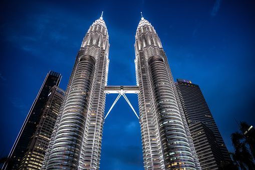 A low-angle shot of two brightly illuminated twin skyscrapers with a sky bridge connecting them against a deep blue night sky.