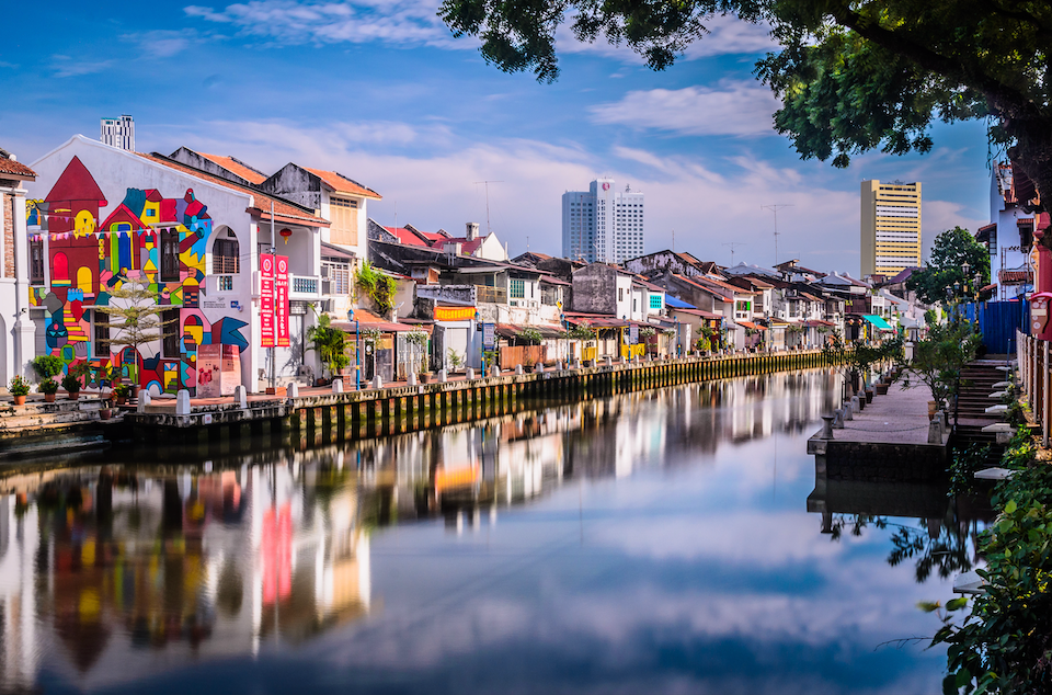 Colorful buildings, one with a large mural, line a calm river, their facades reflected in the water under a blue sky.