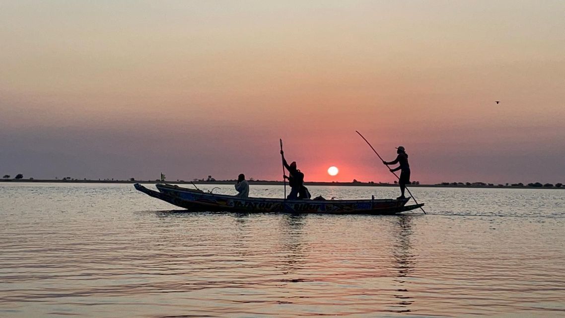 Siluetas de tres personas en un bote en el agua, con dos usando palos mientras el sol se pone en el horizonte.