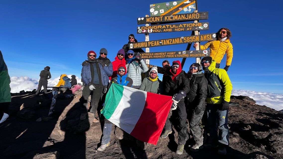 Un gruppo WeRoad in posa con una bandiera italiana davanti a un cartello di legno sulla cima di una montagna, molto al di sopra delle nuvole.