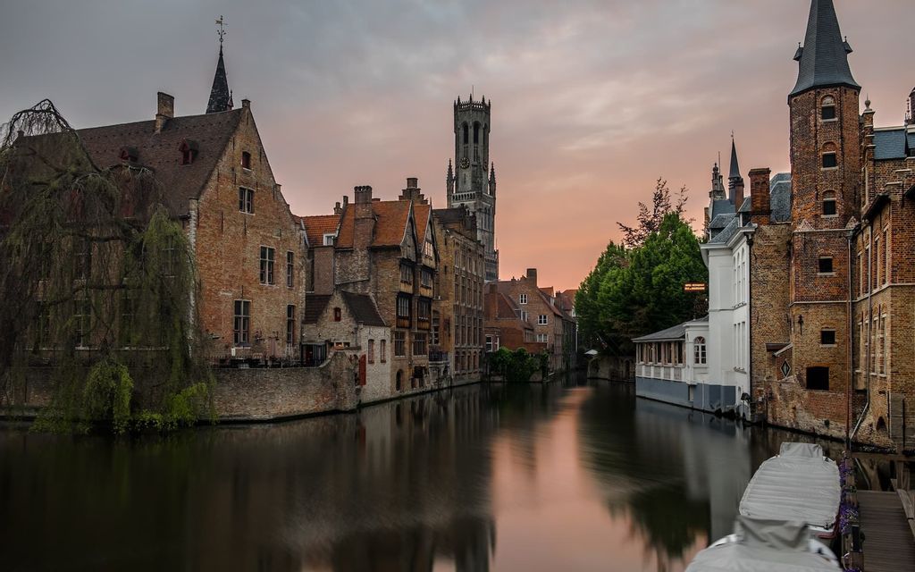 Edificios históricos de ladrillo y una torre del reloj reflejados en un canal urbano al anochecer, con sus imágenes espejadas en el agua.