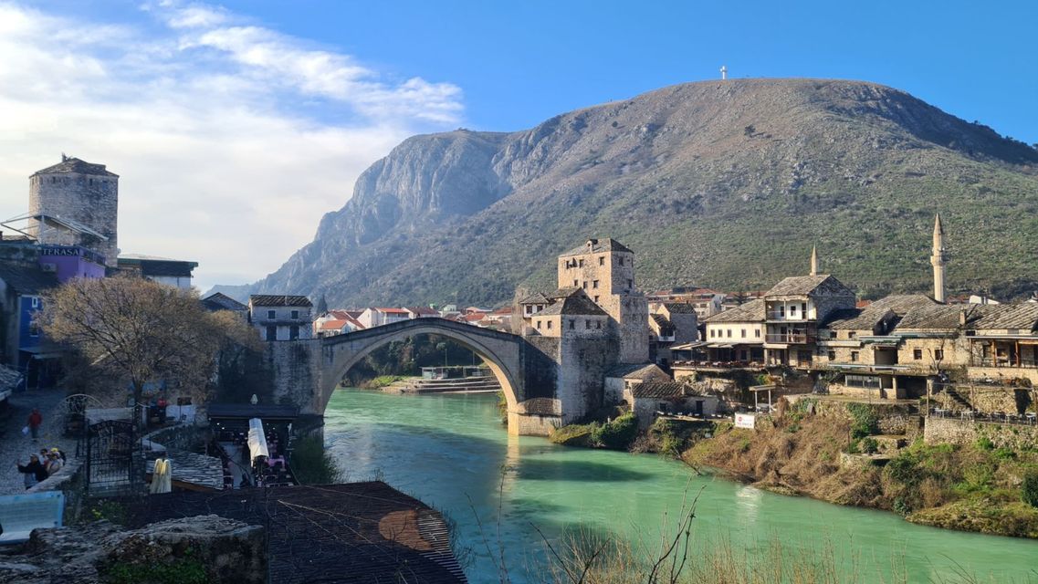 Eine Steinbogenbrücke überspannt einen türkisfarbenen Fluss, der durch eine Stadt mit Steingebäuden und Türmen fließt, mit einem großen Berg im Hintergrund.