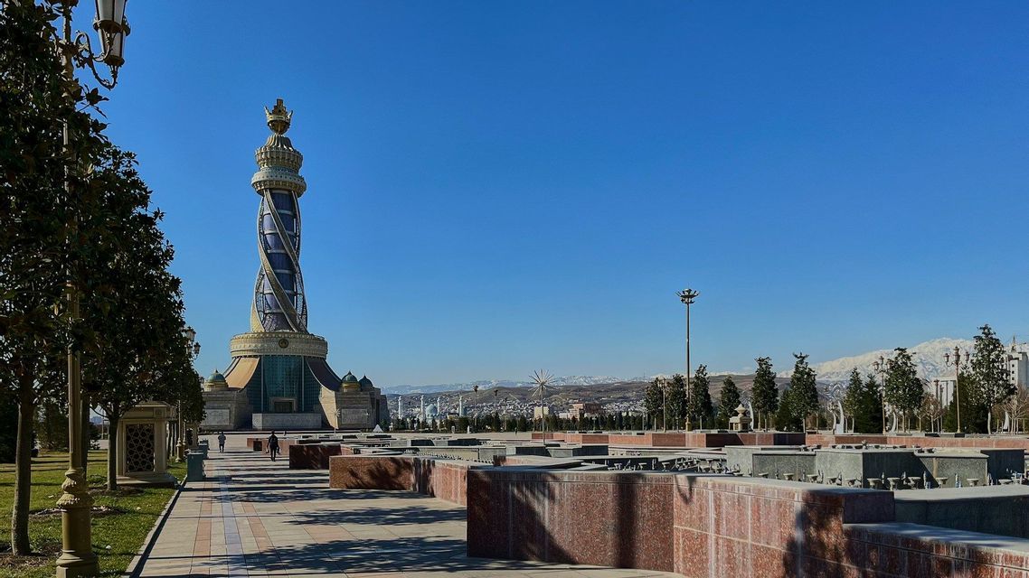 Un monumento ornato e a spirale con una cima dorata si erge in un'ampia piazza, con montagne innevate visibili in lontananza.