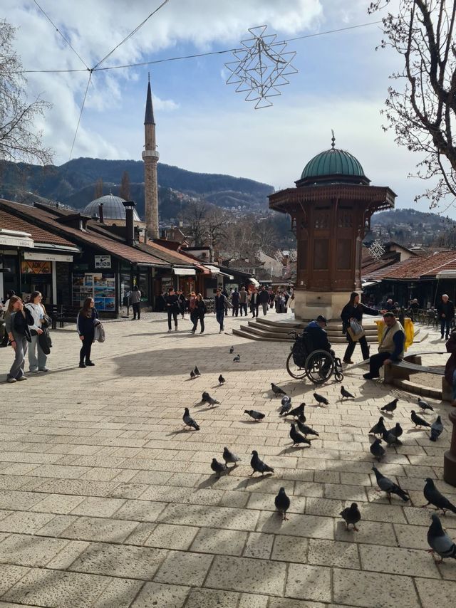Menschen und Tauben versammeln sich auf einem historischen Stadtplatz mit einem markanten Holzbrunnen, einem Minarett und Bergen im Hintergrund.