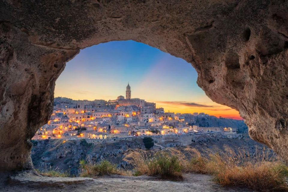 Una città antica e illuminata con un imponente campanile, vista dall'ingresso di una grotta al tramonto.